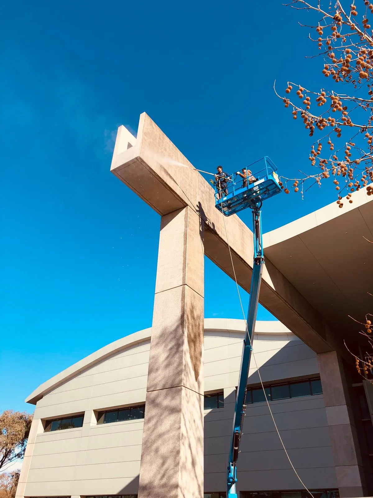 Two workers in a raised boom lift basket pressure-washing the top of a tall concrete blade pylon at a building entrance