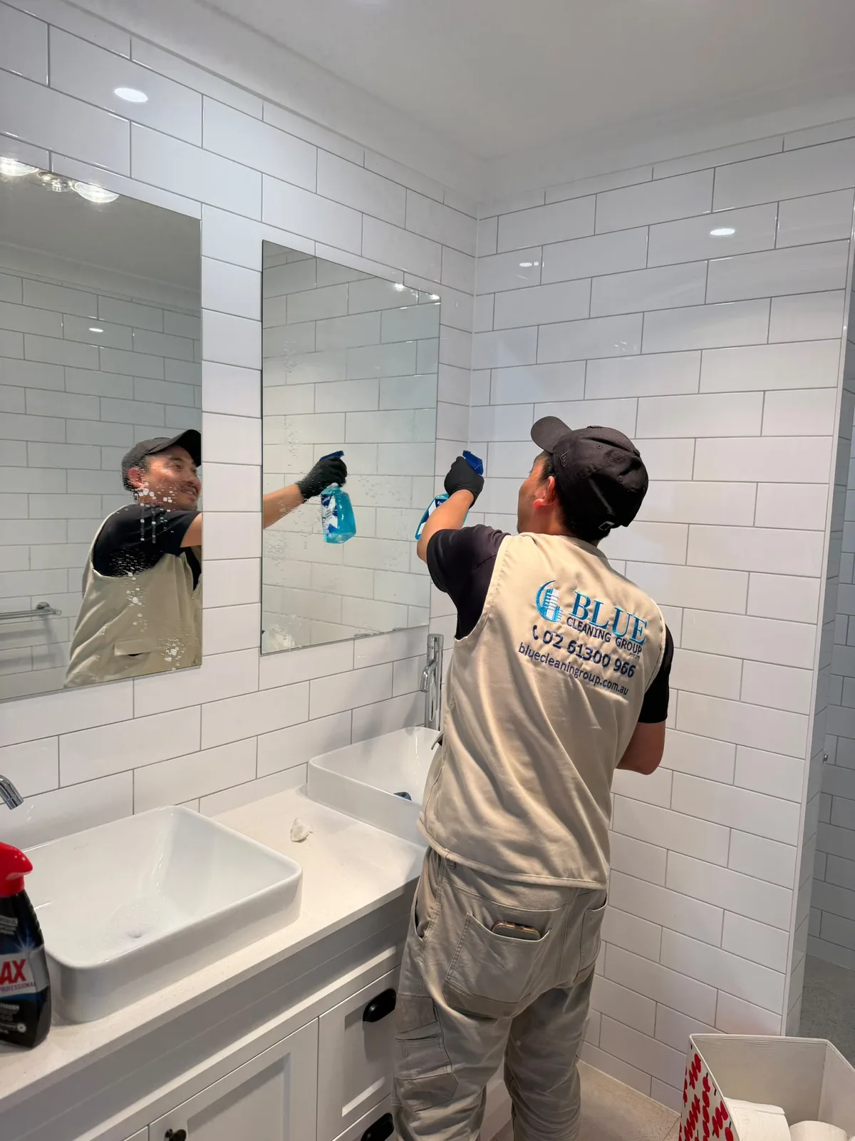 A cleaner in a Blue Cleaning Group tabard spraying and wiping a bathroom mirror above a white vessel basin