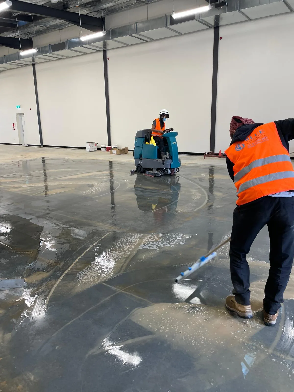 A Blue Cleaning Group worker squeegeeing a flooded slab while a colleague scrubs it on a ride-on machine