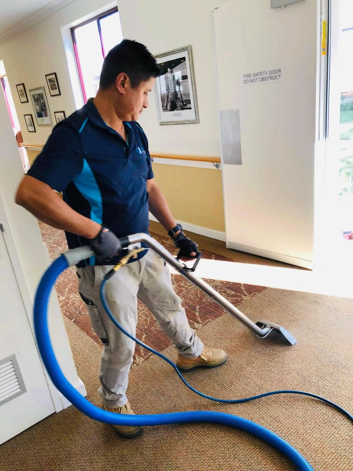 A cleaner working a carpet extraction wand along a corridor beside a stair handrail