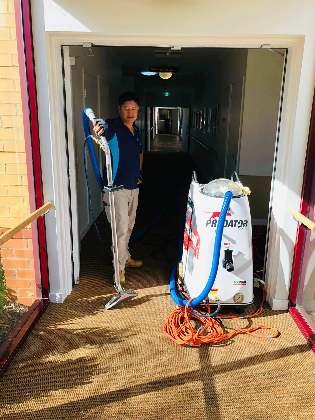 A cleaner drawing a hot water extraction wand back across corridor carpet in a doorway
