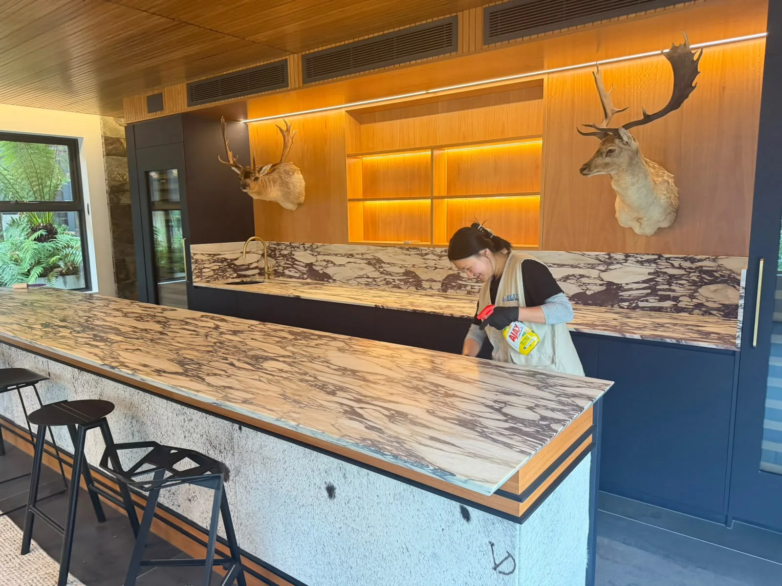 A cleaner leaning across a marble kitchen island with a cloth and a spray bottle, bar stools and navy cabinetry either side