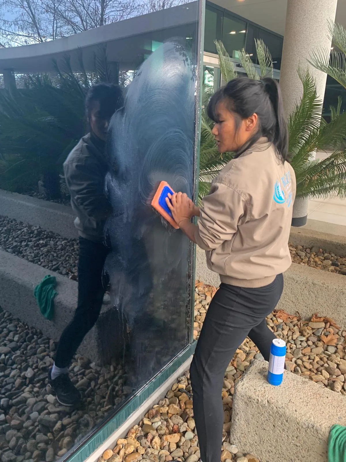 A cleaner in a Blue Cleaning Group jacket working an applicator in circles across a soaped glass panel at an entrance