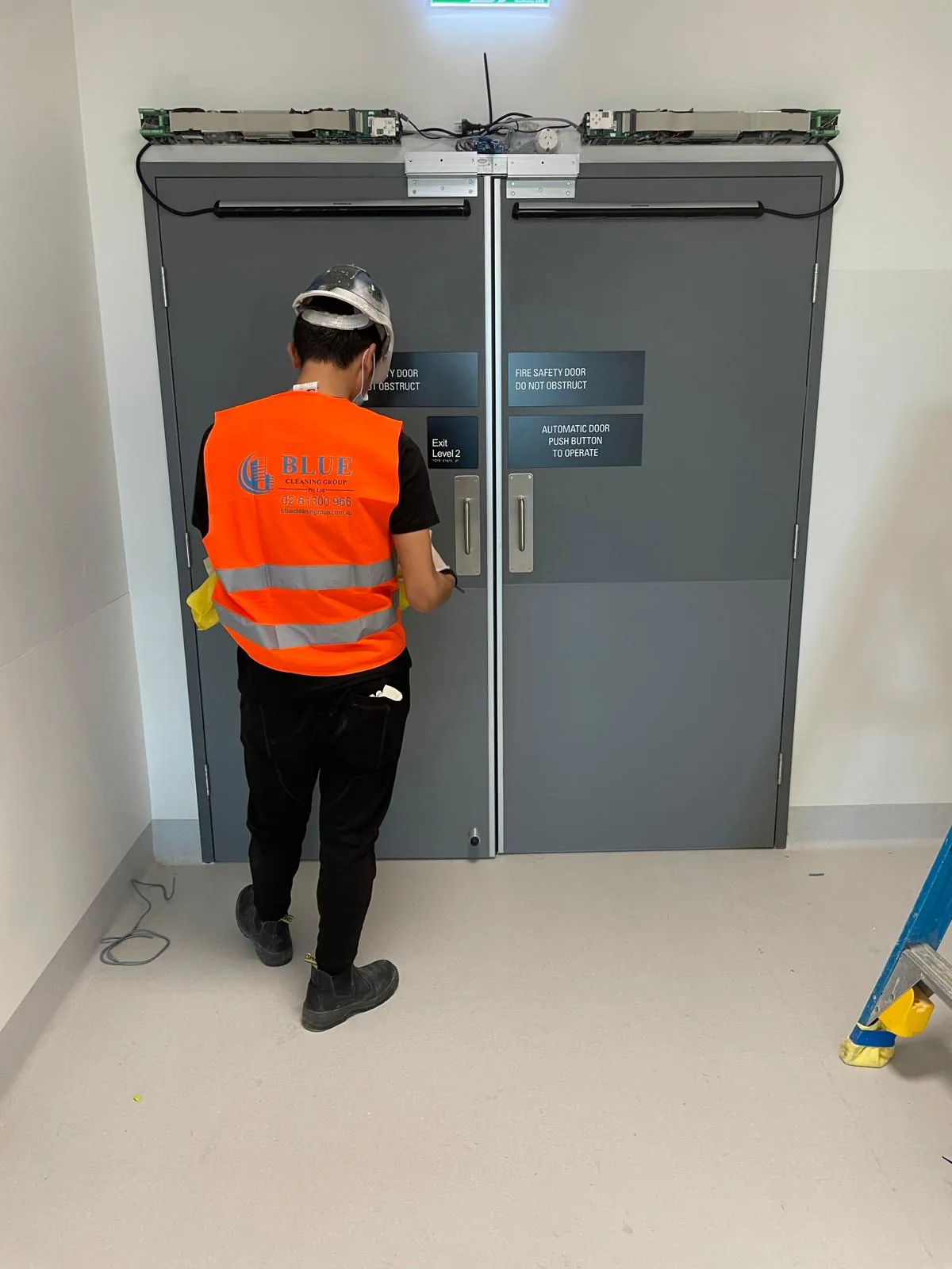 A cleaner in a Blue Cleaning Group vest wiping the handle of a grey fire safety door marked Exit Level 2