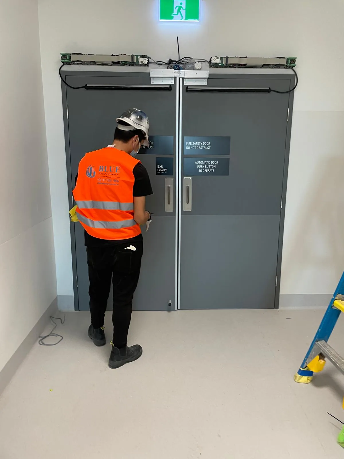 A cleaner in a printed Blue Cleaning Group vest and hard hat wiping the stainless handles of a pair of grey fire safety doors