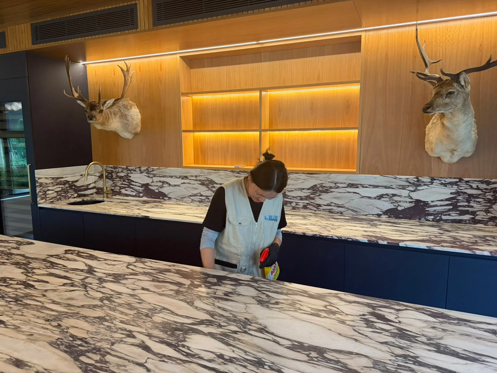A cleaner in a Blue Cleaning Group tabard wiping a marble bar top, the branded tabard legible across her back
