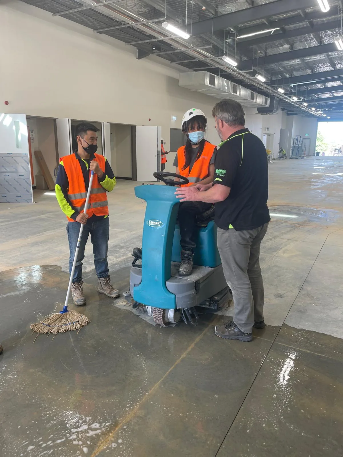 Two cleaners in orange Blue Cleaning Group vests, one leaning on a mop and one seated on a Tennant ride-on scrubber