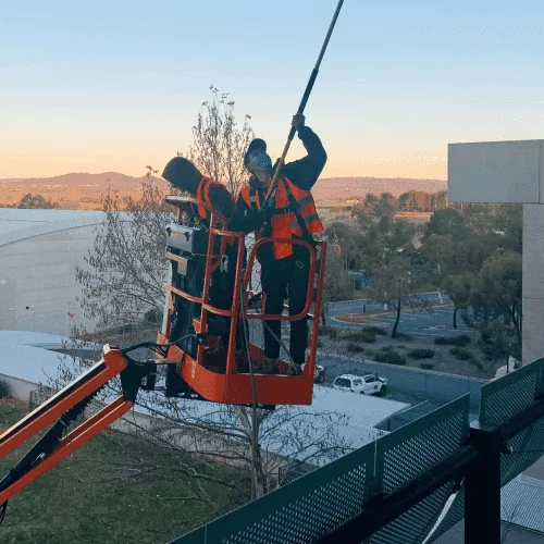 Two workers in a boom lift basket using a pole against a building at sunset over Canberra