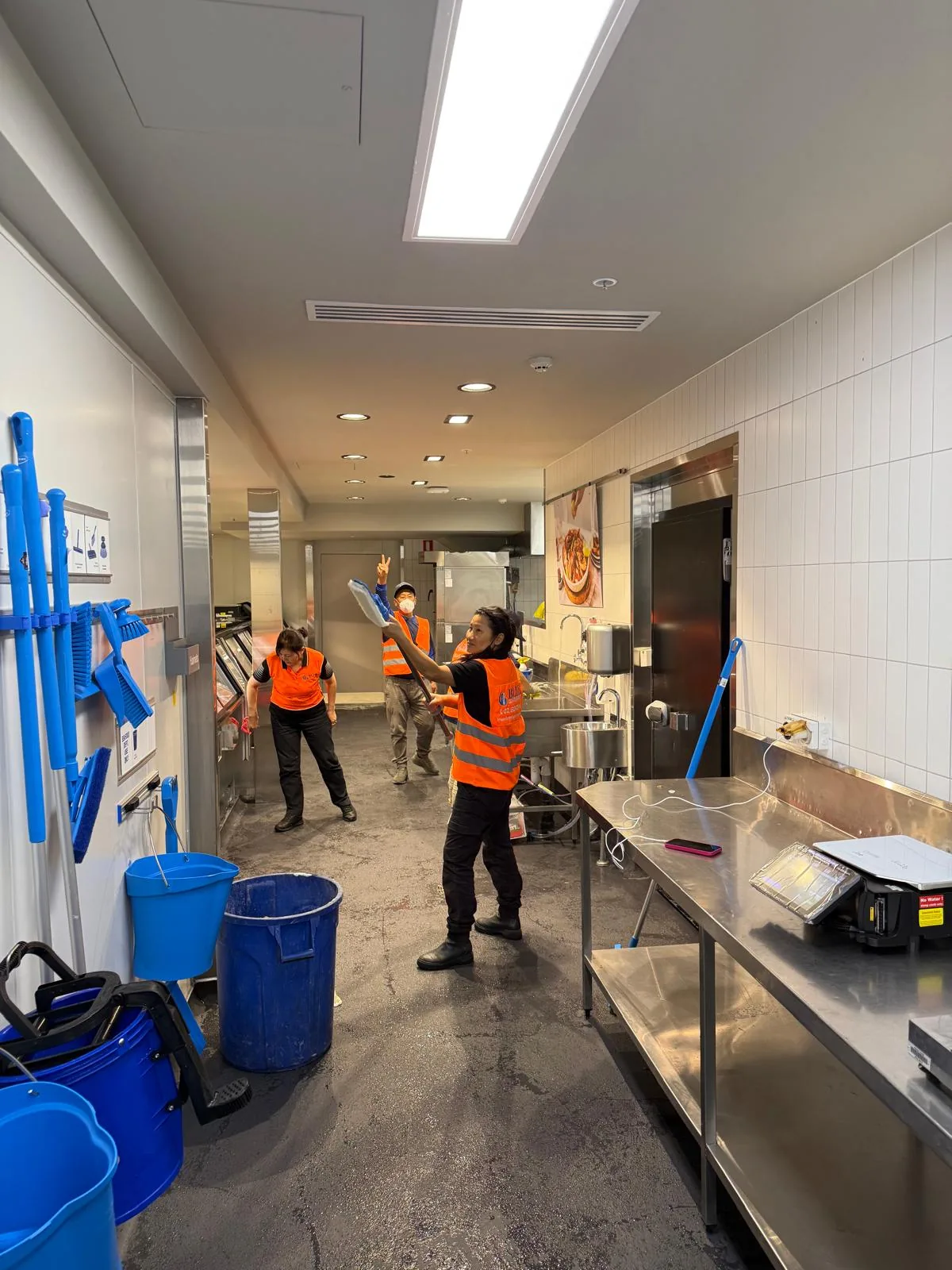 Three workers in orange Blue Cleaning Group vests working a narrow commercial kitchen galley
