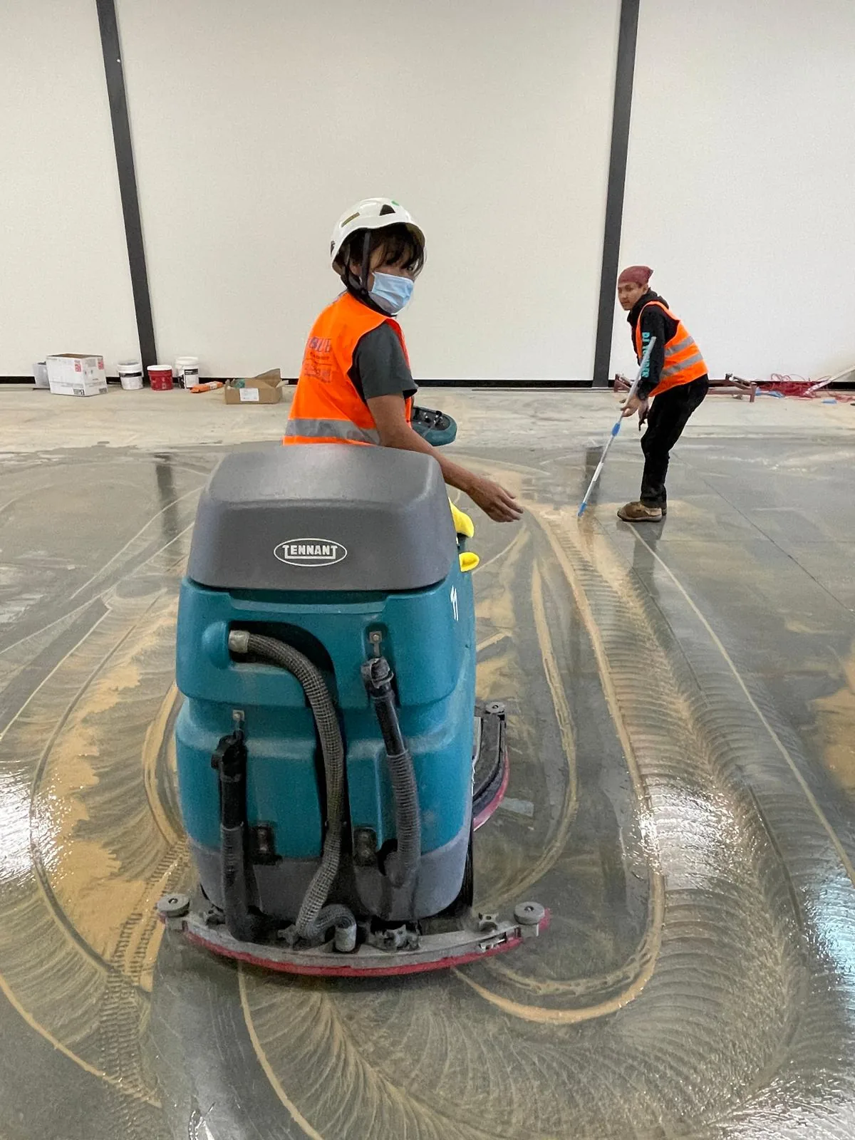 Two cleaners in orange hi-vis vests and hard hats work a flooded concrete slab in an unfinished warehouse