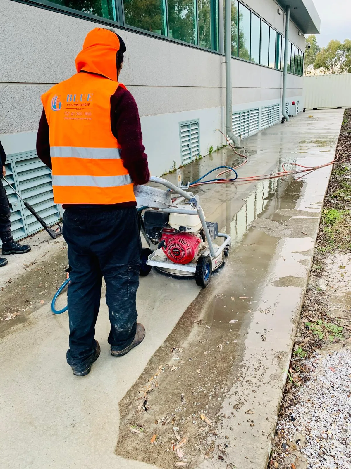Two workers pressure washing a concrete footpath along a commercial building