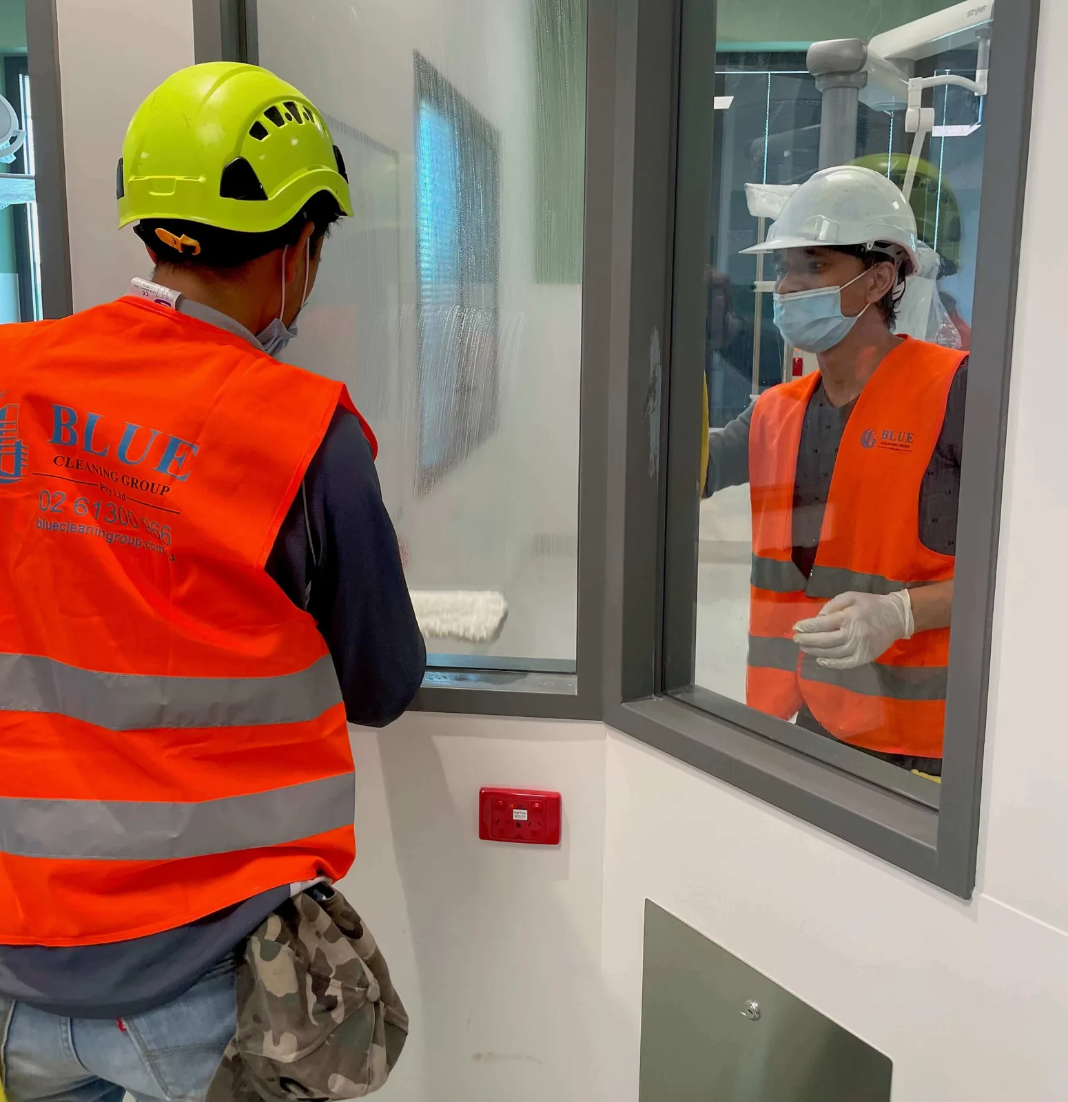 Two cleaners in printed Blue Cleaning Group vests washing an internal window from both sides