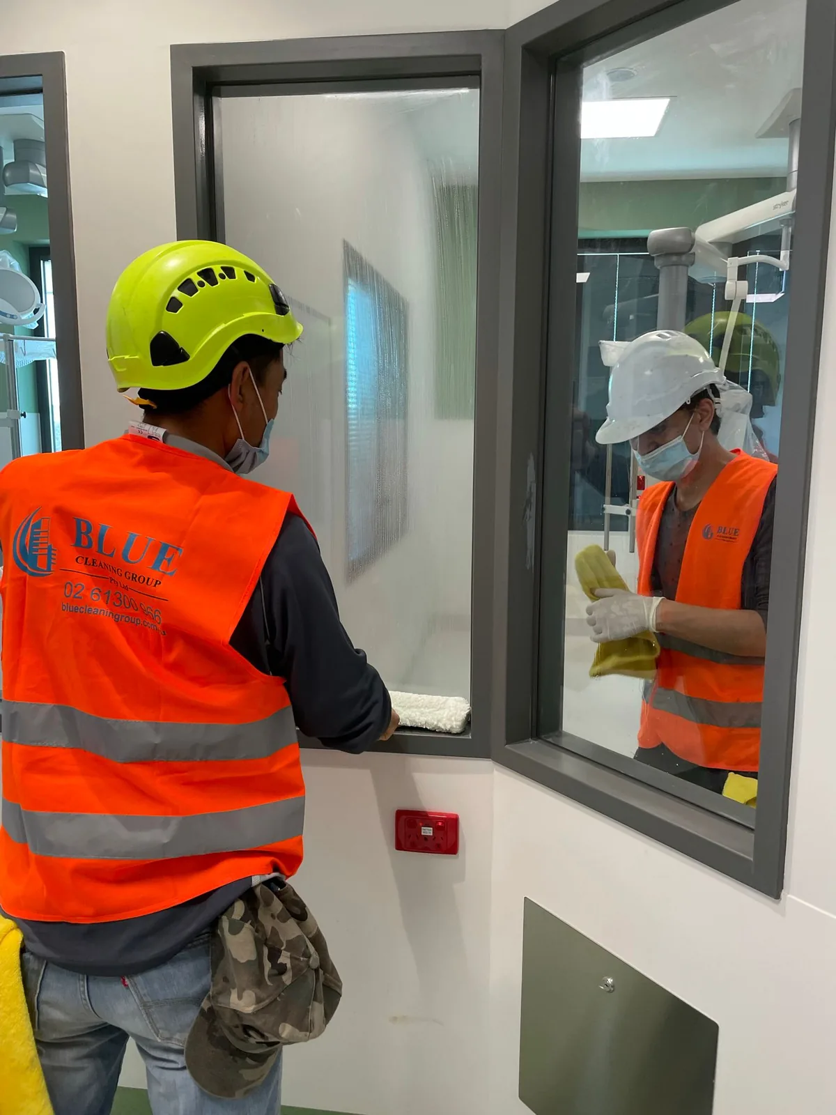Two workers in Blue Cleaning Group hi-vis vests and hard hats cleaning either side of an internal window