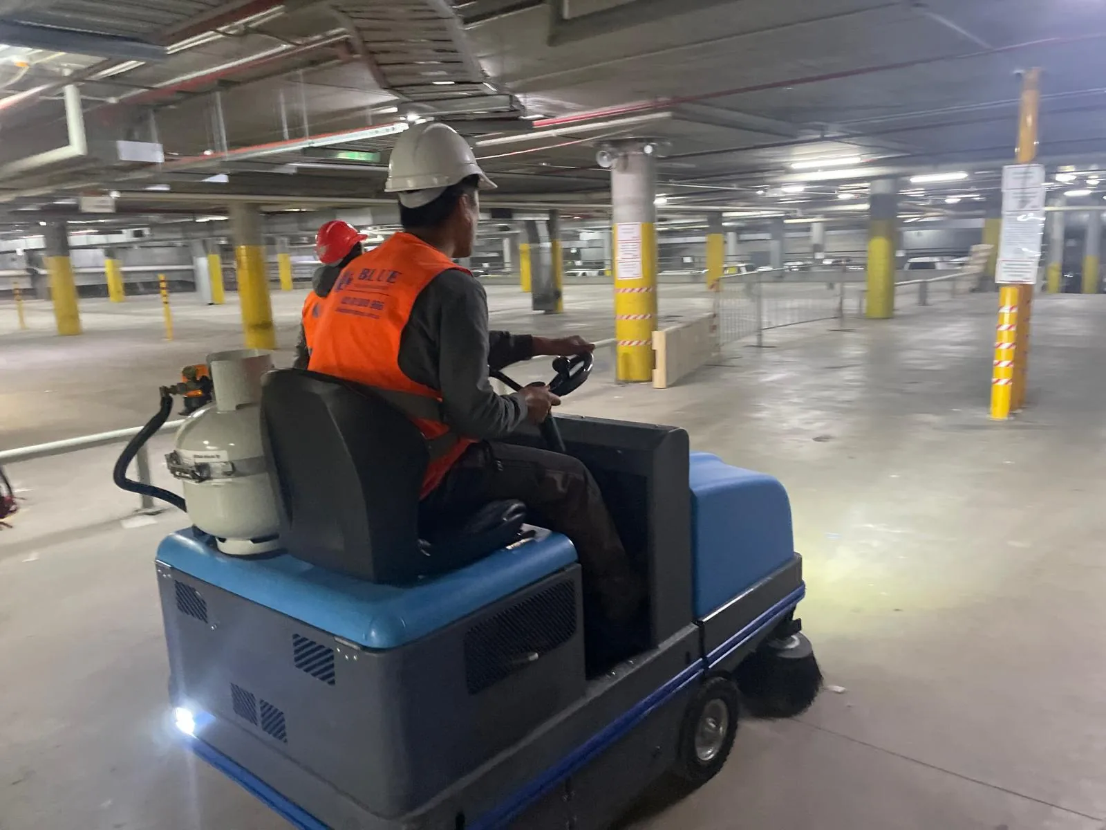 A worker in a hard hat driving an LPG ride-on sweeper across an empty multi-storey car park deck