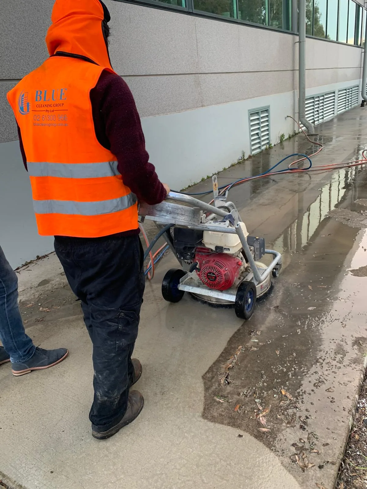 A Blue Cleaning Group worker pushing a rotary surface cleaner along a wet footpath beside a rendered building