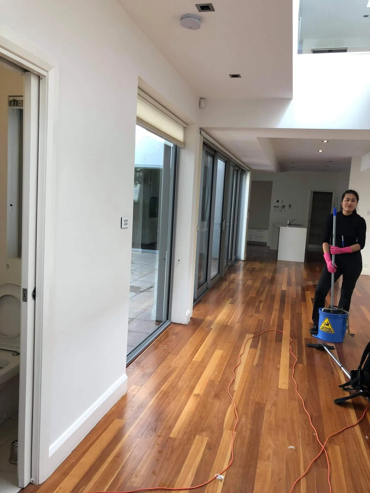 A cleaner mopping the timber floor of an emptied house, a wet-floor bucket beside her and the rooms behind her cleared