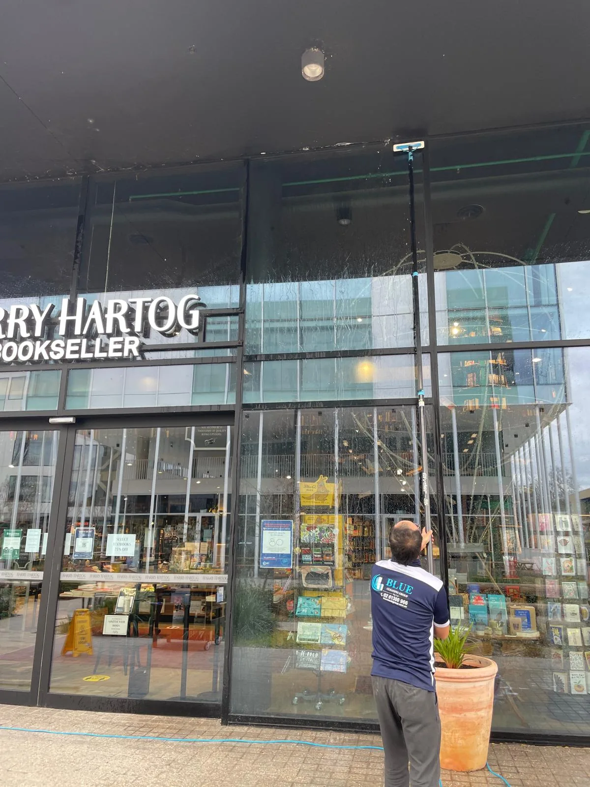 A cleaner in a navy Blue Cleaning Group polo working a water-fed pole up the full-height shopfront glass of a bookshop