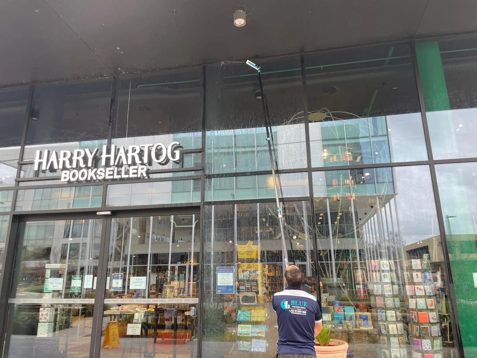 A cleaner in a navy Blue polo running a water-fed pole down a wet pane of a bookshop glass frontage
