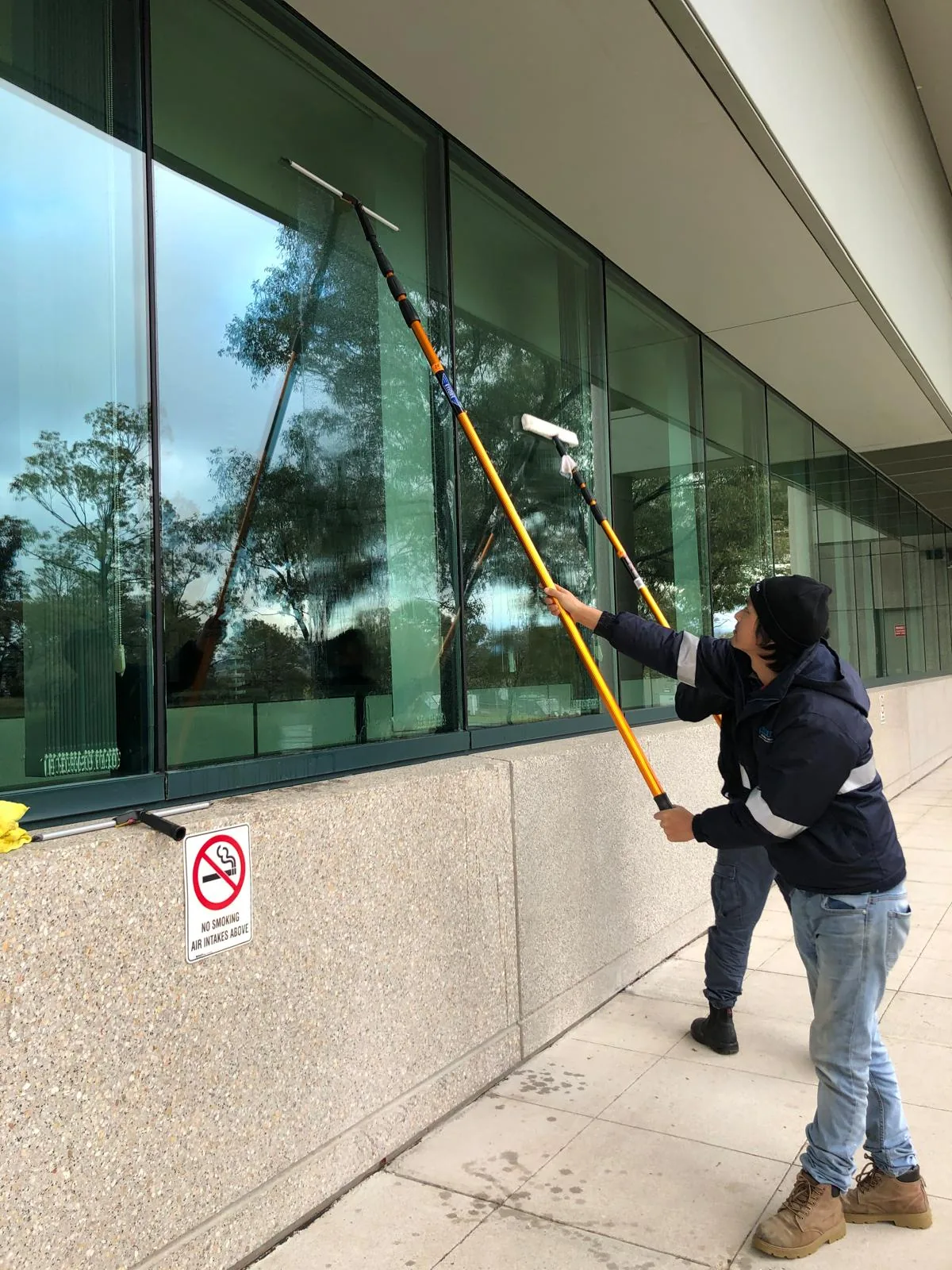 A worker reaching up from the footpath with two extension poles, a washer head and a squeegee
