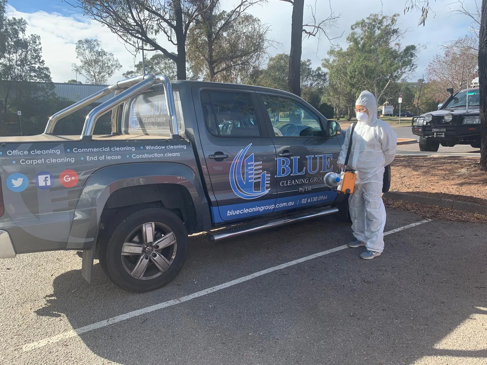 A worker in a coverall and mask holding a thermal fogging machine beside the signwritten Blue Cleaning Group ute