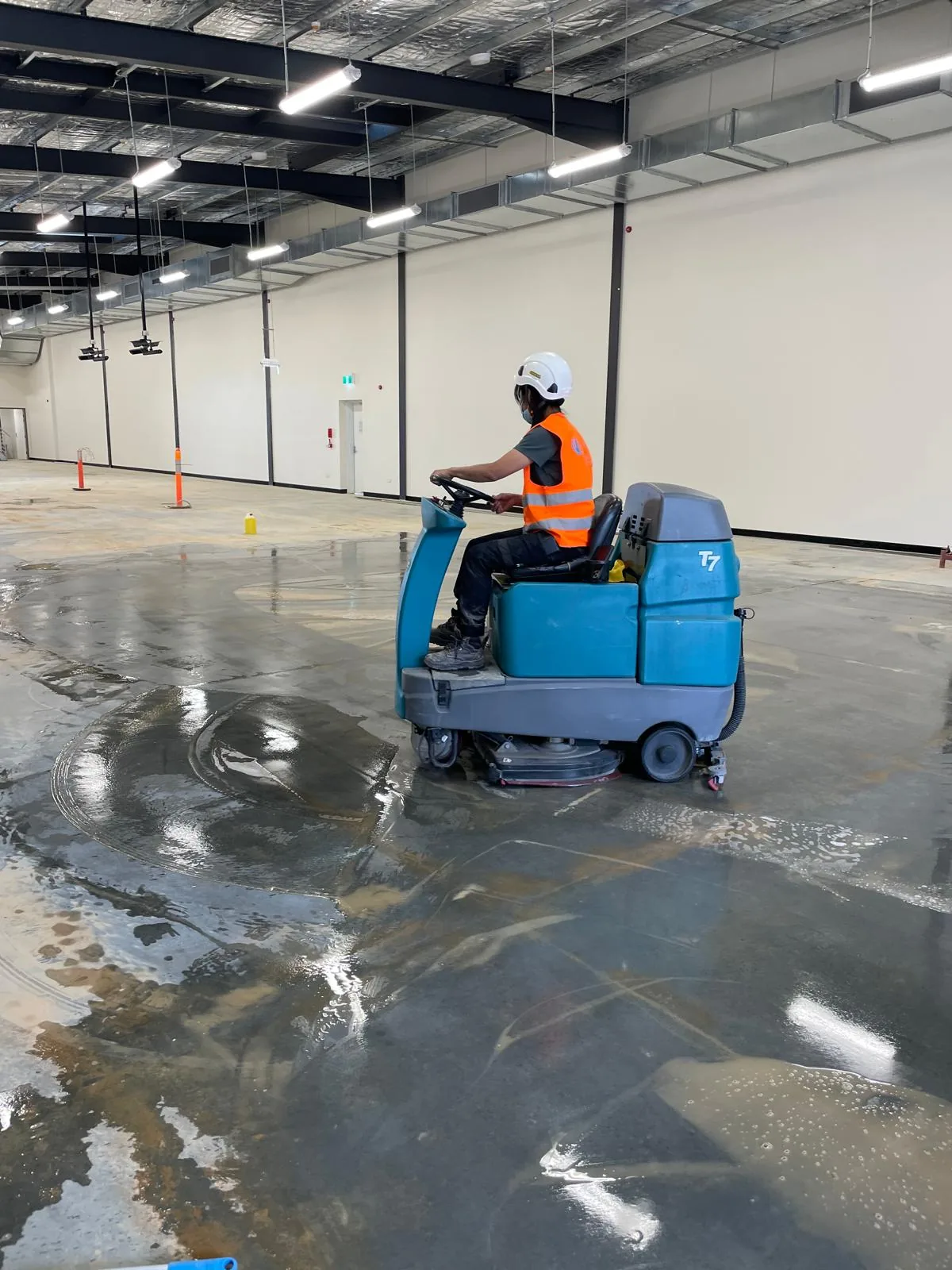 A hard-hatted operator driving a Tennant T7 ride-on scrubber across the wet slab of an empty warehouse shell
