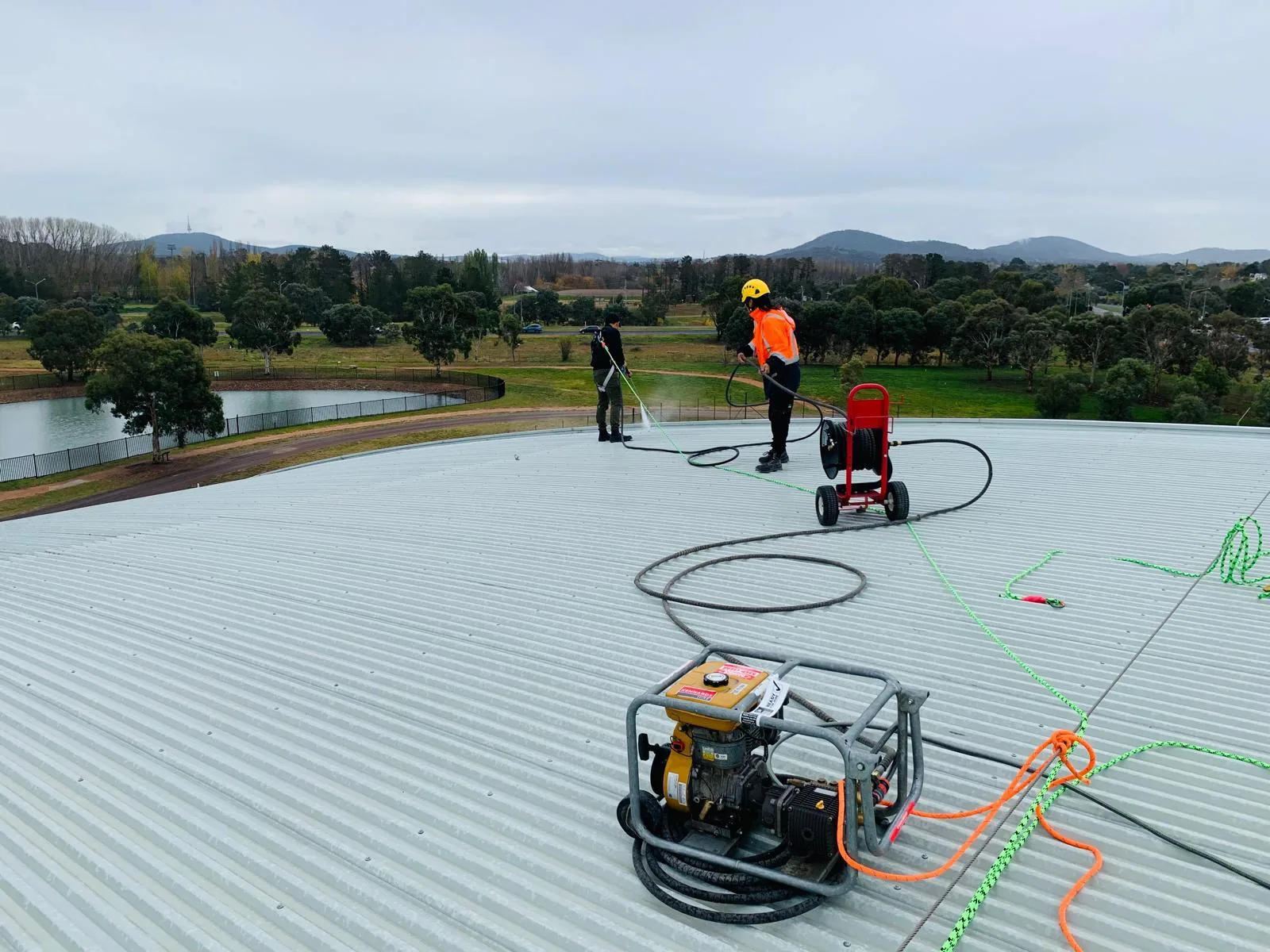 Two harnessed workers washing a large metal roof with lifelines and a hose reel