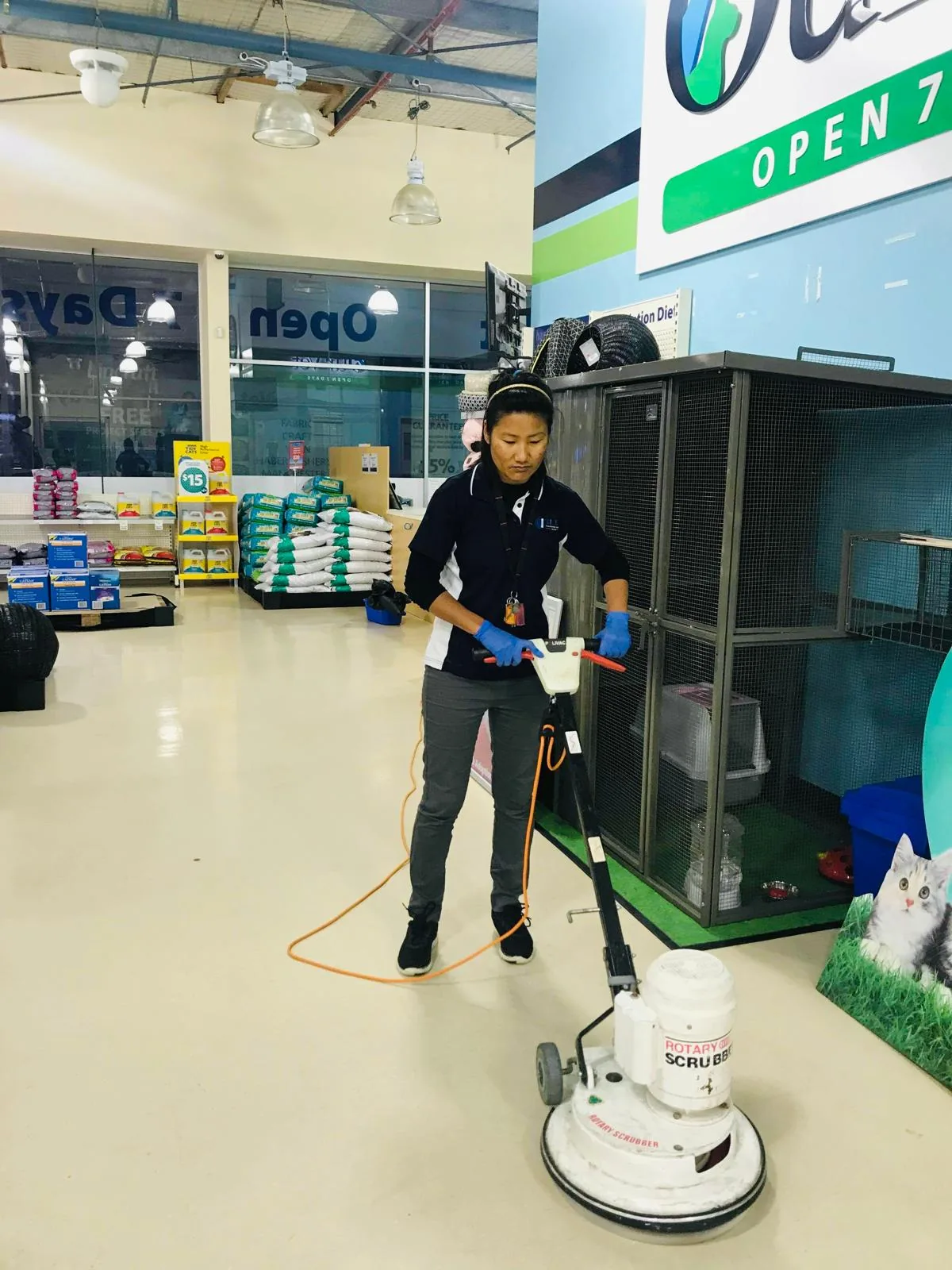 A cleaner in a navy Blue Cleaning Group polo walking a rotary floor scrubber across the vinyl floor of a shop