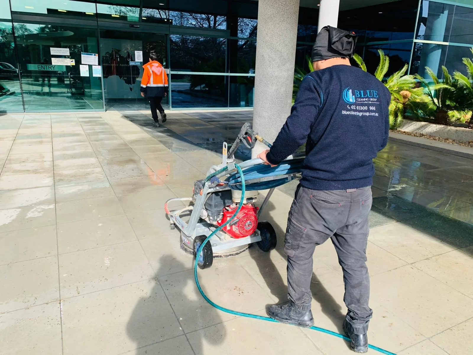 A worker running a petrol rotary surface cleaner across an office forecourt