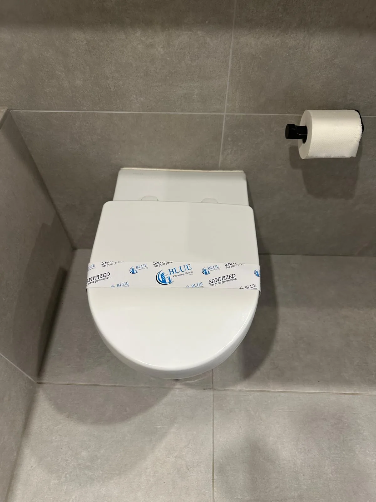 A Blue Cleaning Group sanitised band across a closed toilet lid in a tiled bathroom