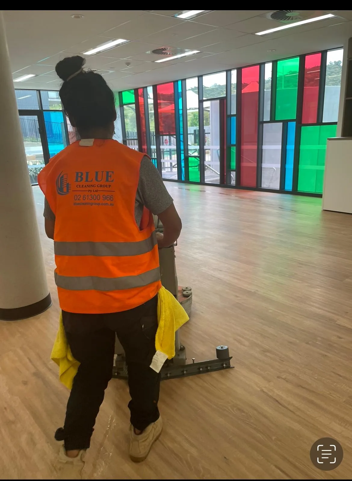 A Blue Cleaning Group cleaner pushing a floor scrubber across a large unfurnished hall glazed with coloured panels