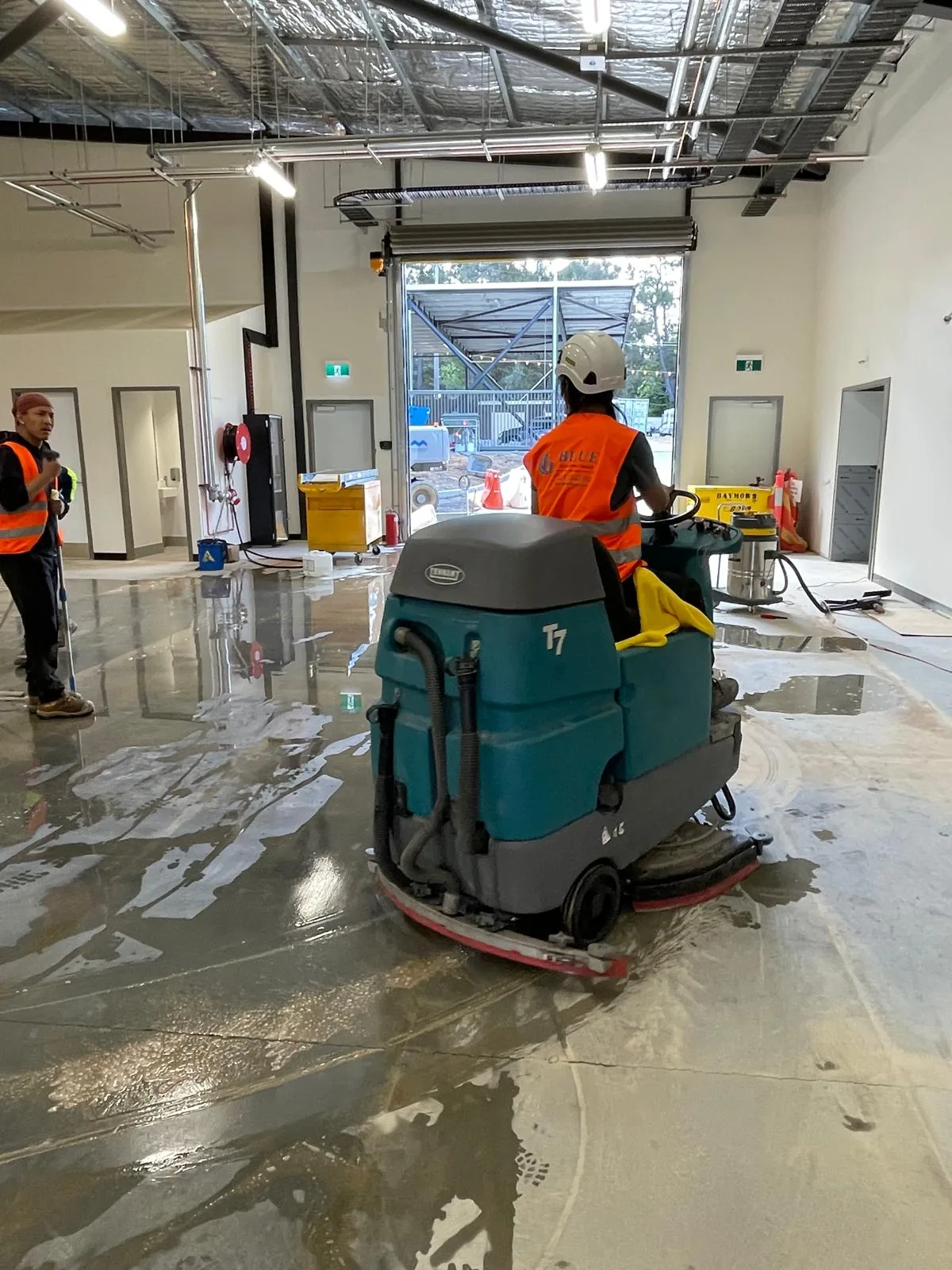 A Blue Cleaning Group operator driving a Tennant T7 ride-on scrubber over a wet slab towards an open roller door