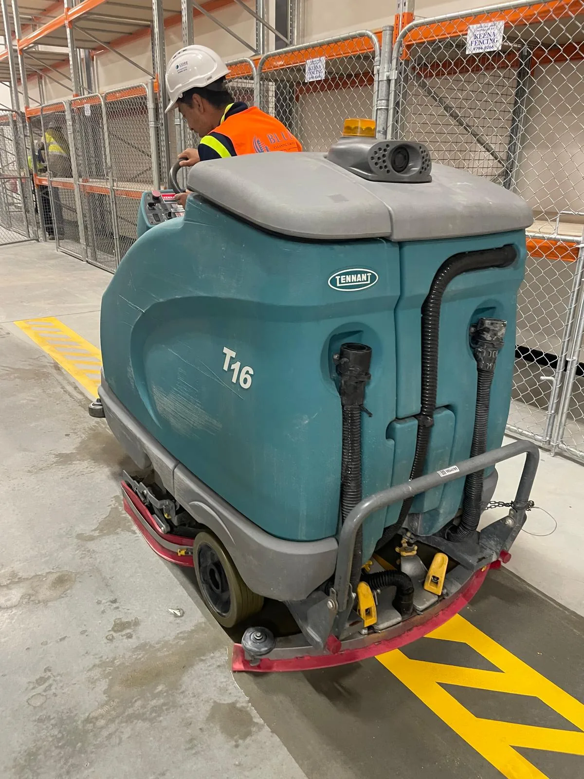 A Blue Cleaning operator in a hard hat driving a Tennant T16 scrubber down a racking aisle in a distribution warehouse