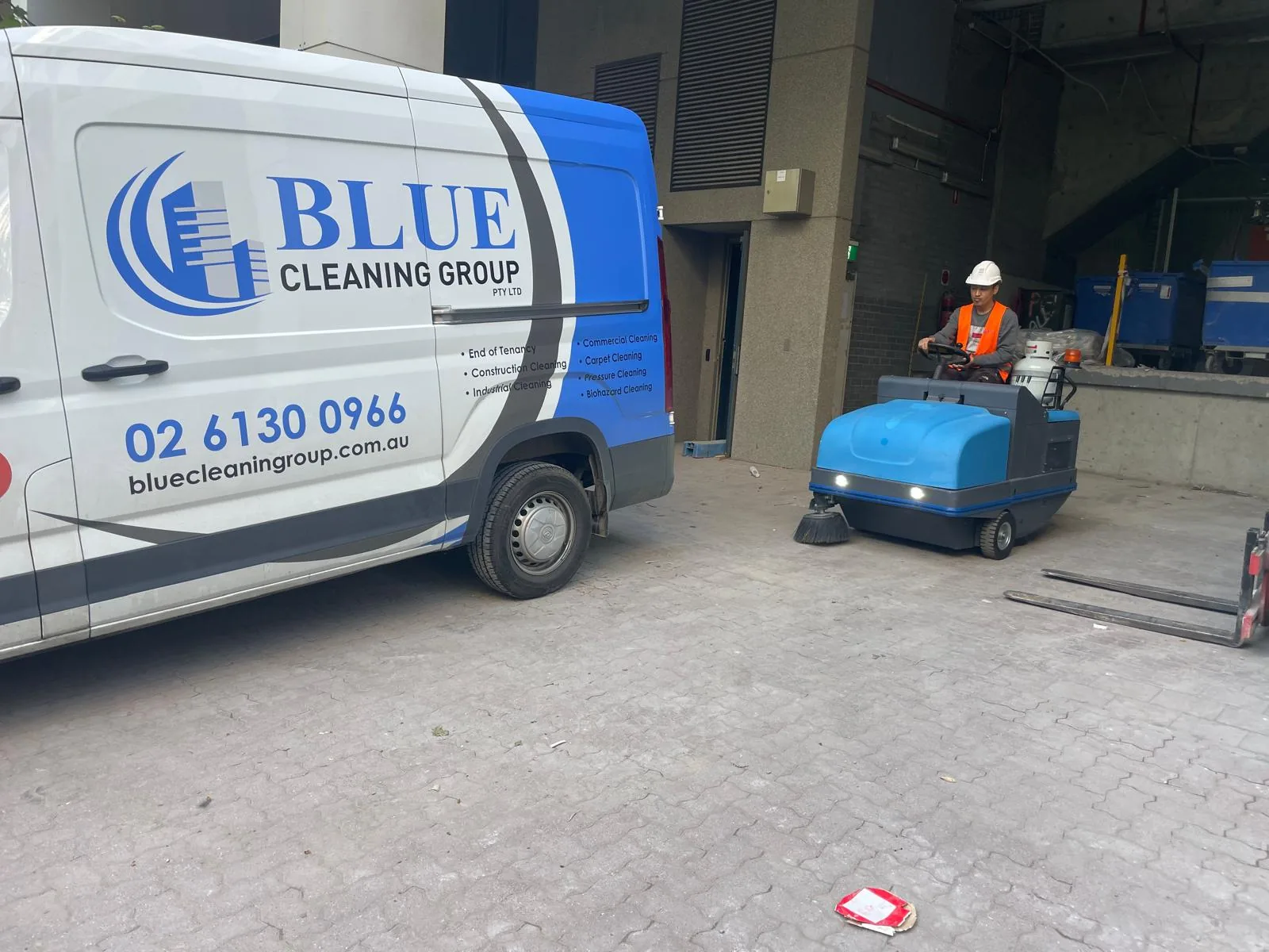 The signwritten Blue Cleaning Group van on a loading bay, a worker driving a ride-on sweeper across the pavers