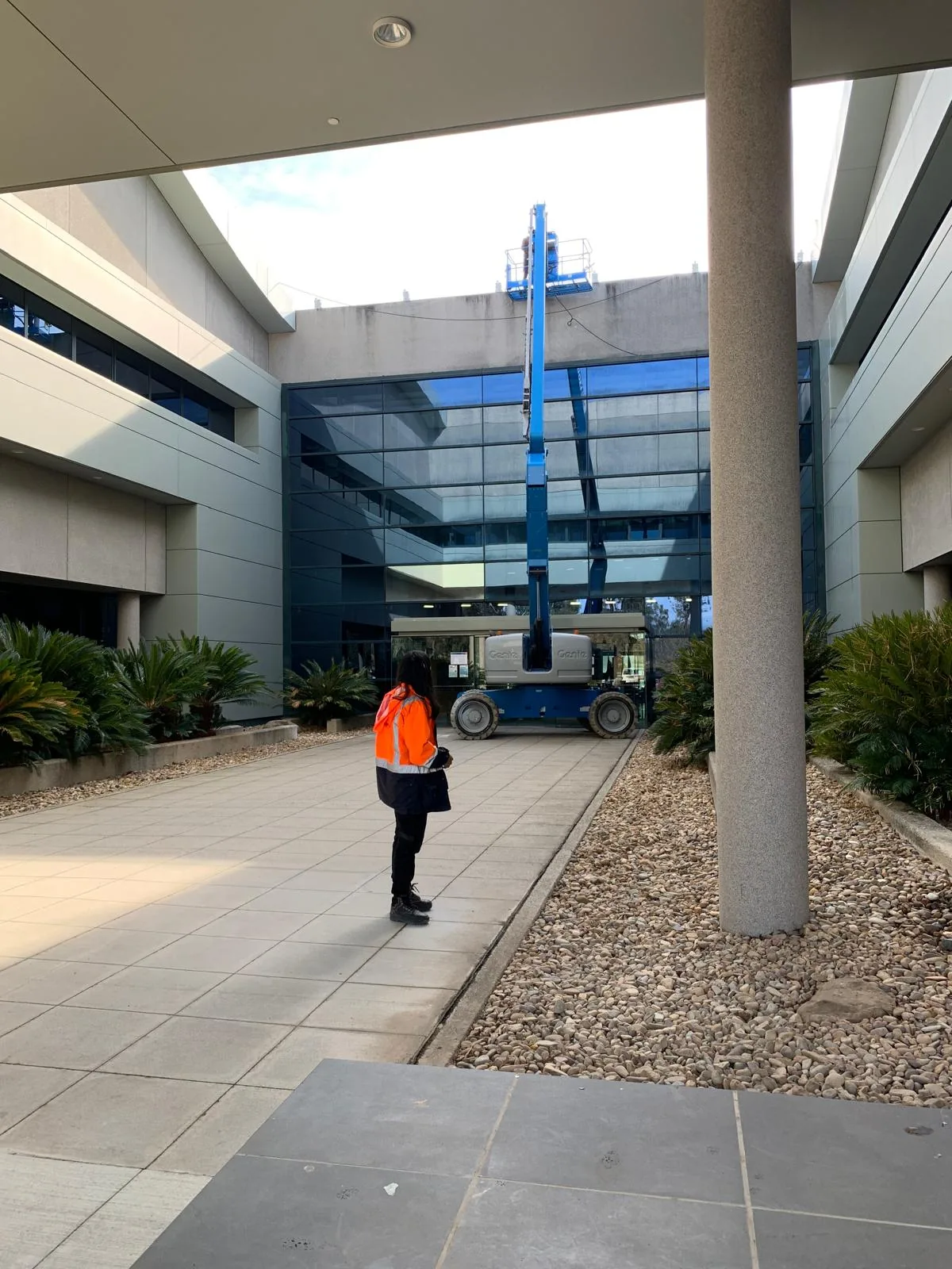 A worker in hi-vis standing in a paved courtyard watching a boom lift raised over the parapet of a glazed office building