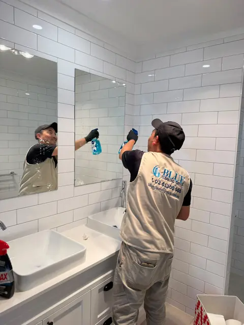 A cleaner in a Blue Cleaning Group tabard wiping a bathroom mirror above a white basin