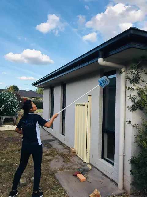 A Blue Cleaning Group cleaner raising a cobweb duster on a pole to the eaves of a single-storey house