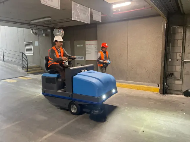 A worker in a hard hat driving a blue ride-on sweeper across a basement car park deck