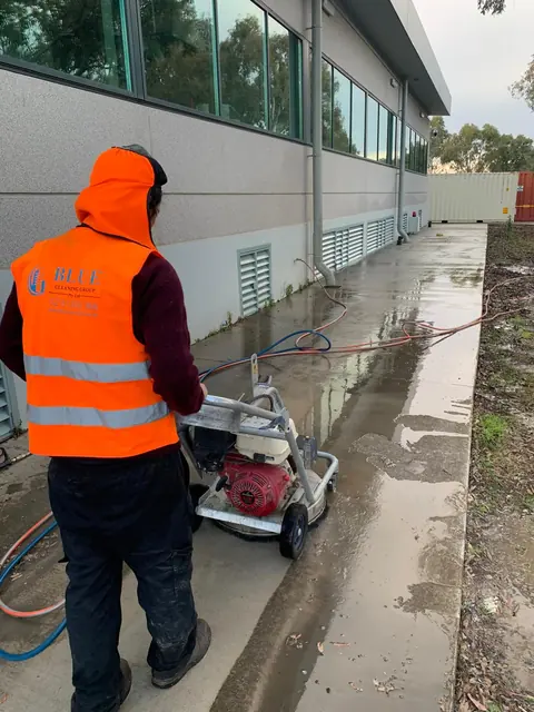 A Blue Cleaning Group worker pushing a rotary surface cleaner down a long wet path beside a commercial building