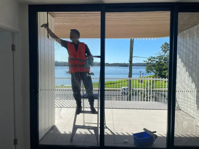 A cleaner on a stepladder washing a balcony sliding door from outside, a lake visible beyond