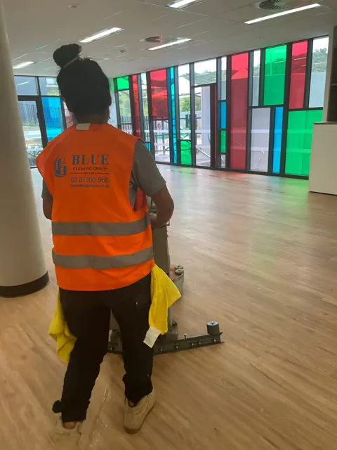 A worker running a rotary floor machine through a hall glazed with coloured panels
