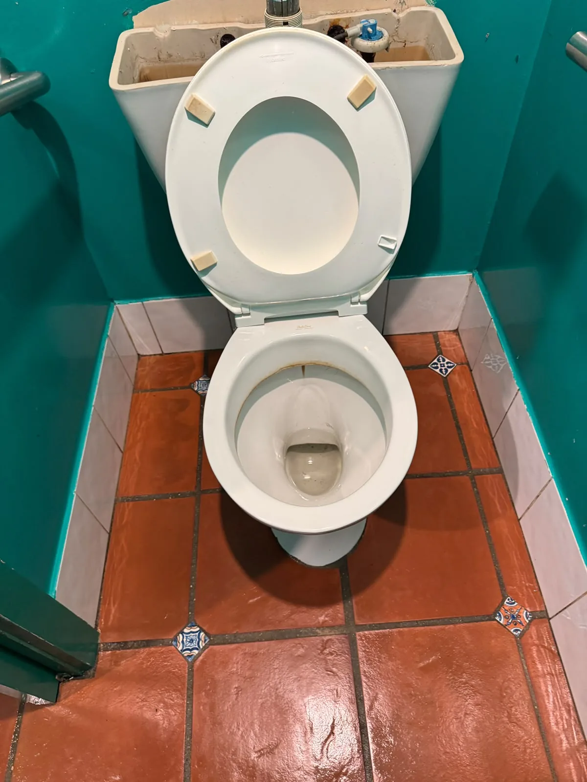 The same teal-walled cubicle cleaned: the toilet white with the seat raised, the terracotta floor tiles washed and still wet