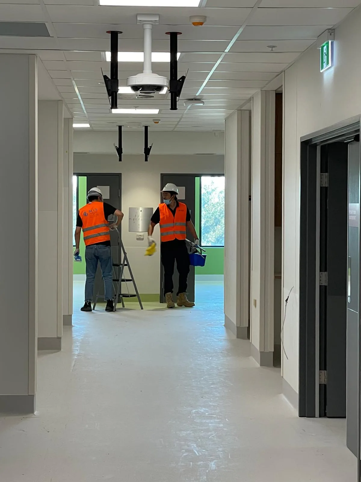 Two cleaners in hard hats and hi-vis detailing doorways in a new corridor, one working from a step ladder