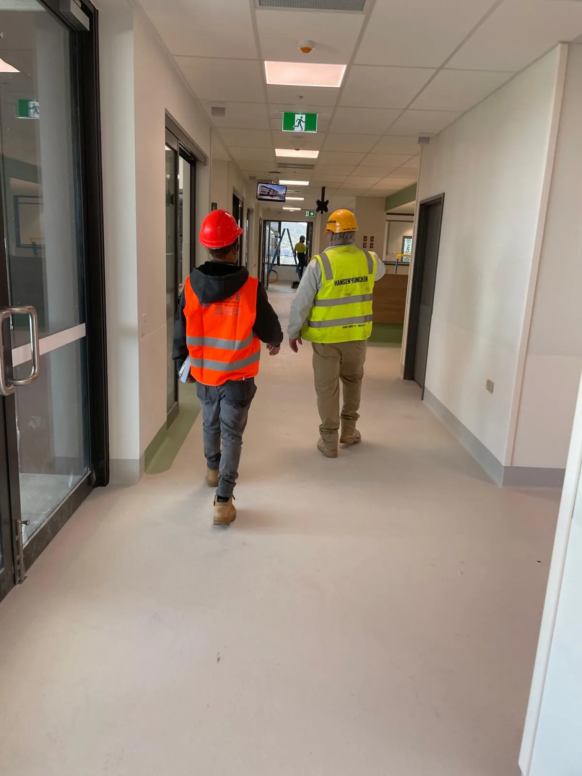 A Blue Cleaning worker in orange hi-vis walking a new hospital corridor beside a builder in a Hansen Yuncken vest